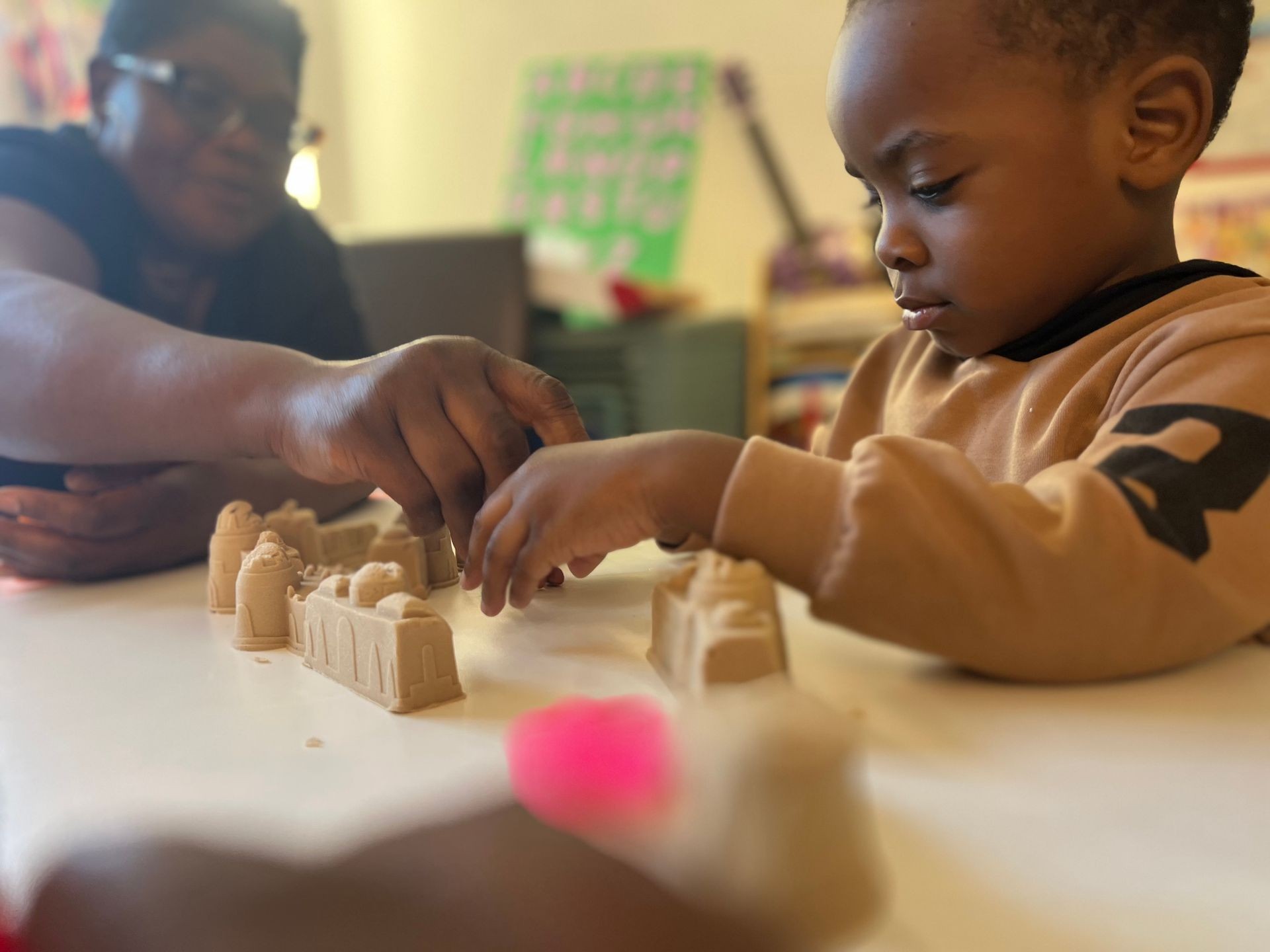 Two people building with sandcastle molds on a table in a brightly lit room with colorful decorations.